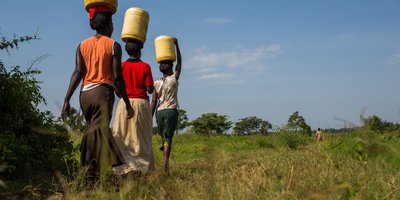 Three women carry water barrels on their heads as they walk through a grassy field.
