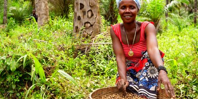 A woman kneels beside a basket full of food in the forest.