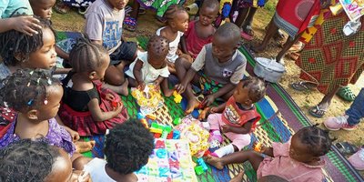 Children of different ages sit in a circle on a blanket playing with toys.