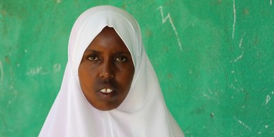 A woman wearing a head scarf stands in front of a green chalkboard.
