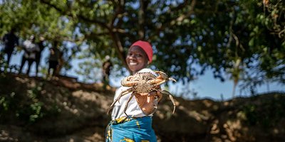 A woman stand in a forest clearing, holding a crab towards the camera.
