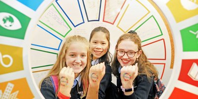 Three schoolchildren hold up their hands to show colourful transfers. Surrounding them is a circular multi-coloured arch.