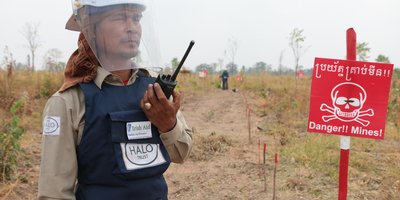 A man wearing a helmet with a plastic face screen, and holding a walkie talkie, looks off camera. Beside him is a red sign with a skull and crossbones and the text: Danger! Mines!