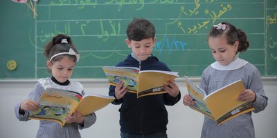Three children stand in front of a green chalk board reading schoolbooks.