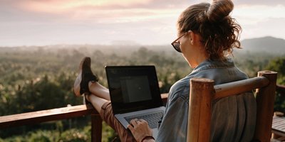 Woman sat and working on her laptop outdoors at sunrise. Her feet are up on the railing