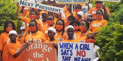 A large group of women wearing orange t-shirts and polo shirts hold placards against violence against women.