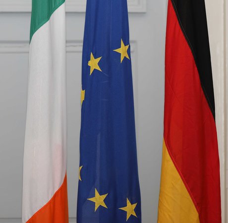 The Irish flag, EU flag and German flag standing side-by-side on indoor flagpoles.