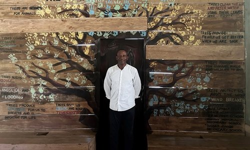 Tommy Garnett stood in a white shirt smiling in front of a mural titled The Importance of Trees that is composed of children's handprints