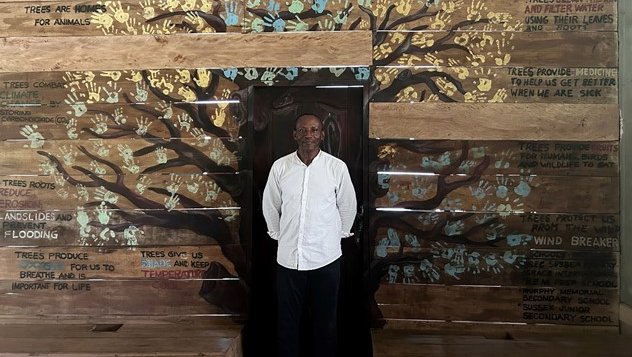Tommy Garnett stood in a white shirt smiling in front of a mural titled The Importance of Trees that is composed of children's handprints