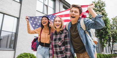 Three young people wearing school bags holding an American flag behind them and laughing together