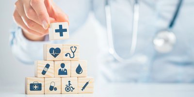 Someone with a stethoscope around their neck placing a block with the first aid symbol on a pyramid of blocks with various medical symbols on them