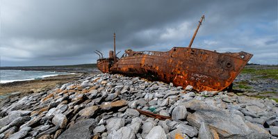A rocky beach with a blurred view of a rusted shipwreck in the background.