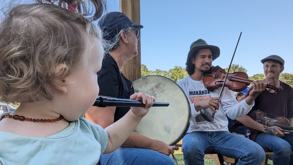 A young girl playing the tin whistle along with adults playing the bodhran and the fiddle.