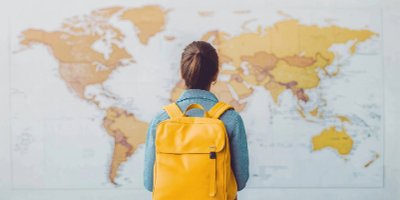 A woman wearing blue and a bright yellow backpack has her back to the camera and is looking at a large map