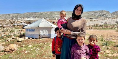 A woman holding a baby is surrounded by three young children. A white UNHCR tent is in the background on a sunny, arid landscape.