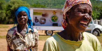 A woman laughs in the foreground as another woman behind her smiles at the camera. In the background is a white truck and a tall green mountain.