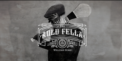 Man with a hurl slung over his shoulder wearing a peat cap with the Auld Fella logo in front of him