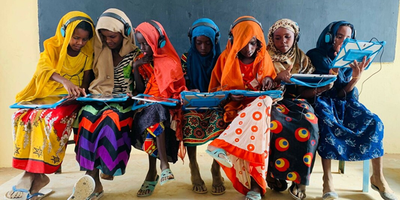 Seven girls in multi-coloured clothing sit in a row, holding tablets and looking at their own or the others' screens.